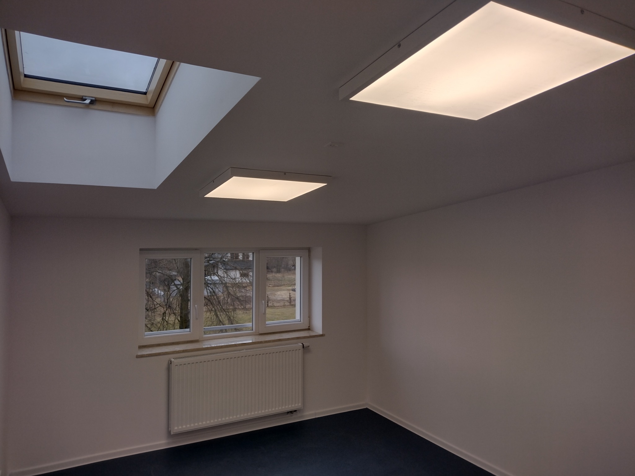 Pusty pokój z oknem dachowym, trzema oknami z widokiem na drzewa i dwoma kwadratowymi lampami sufitowymi LED.