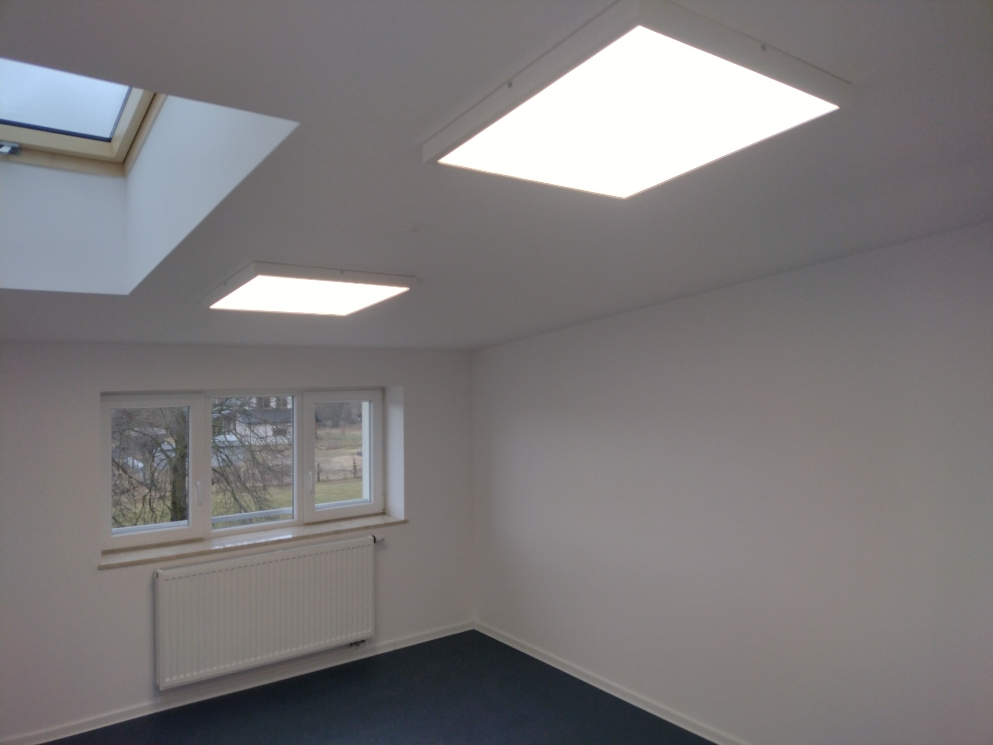 Pomalowane na biało wnętrze biura z oknem, grzejnikiem i dwoma panelami LED w suficie oraz oknem dachowym. Podłoga pokryta ciemną wykładziną.