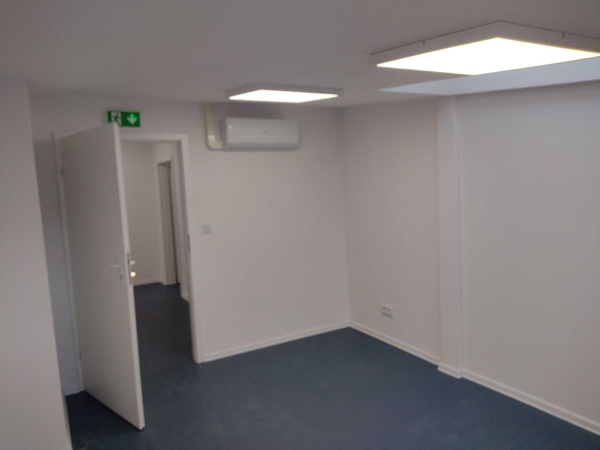 Puste, białe biuro z niebieską podłogą, oświetleniem sufitowym LED, klimatyzacją i otwartymi drzwiami z zielonym znakiem ewakuacyjnym.