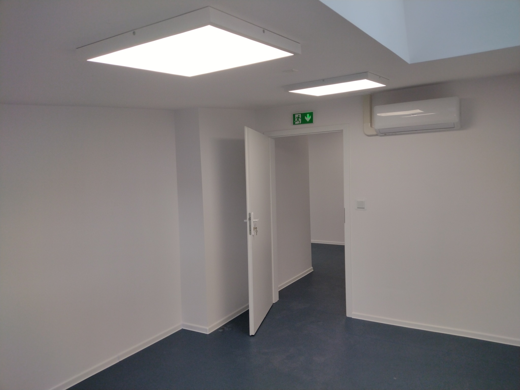 Pomalowane na biało wnętrze biura z otwartymi drzwiami, widoczne lampy sufitowe i klimatyzator. Podłoga wyłożona ciemnym linoleum.