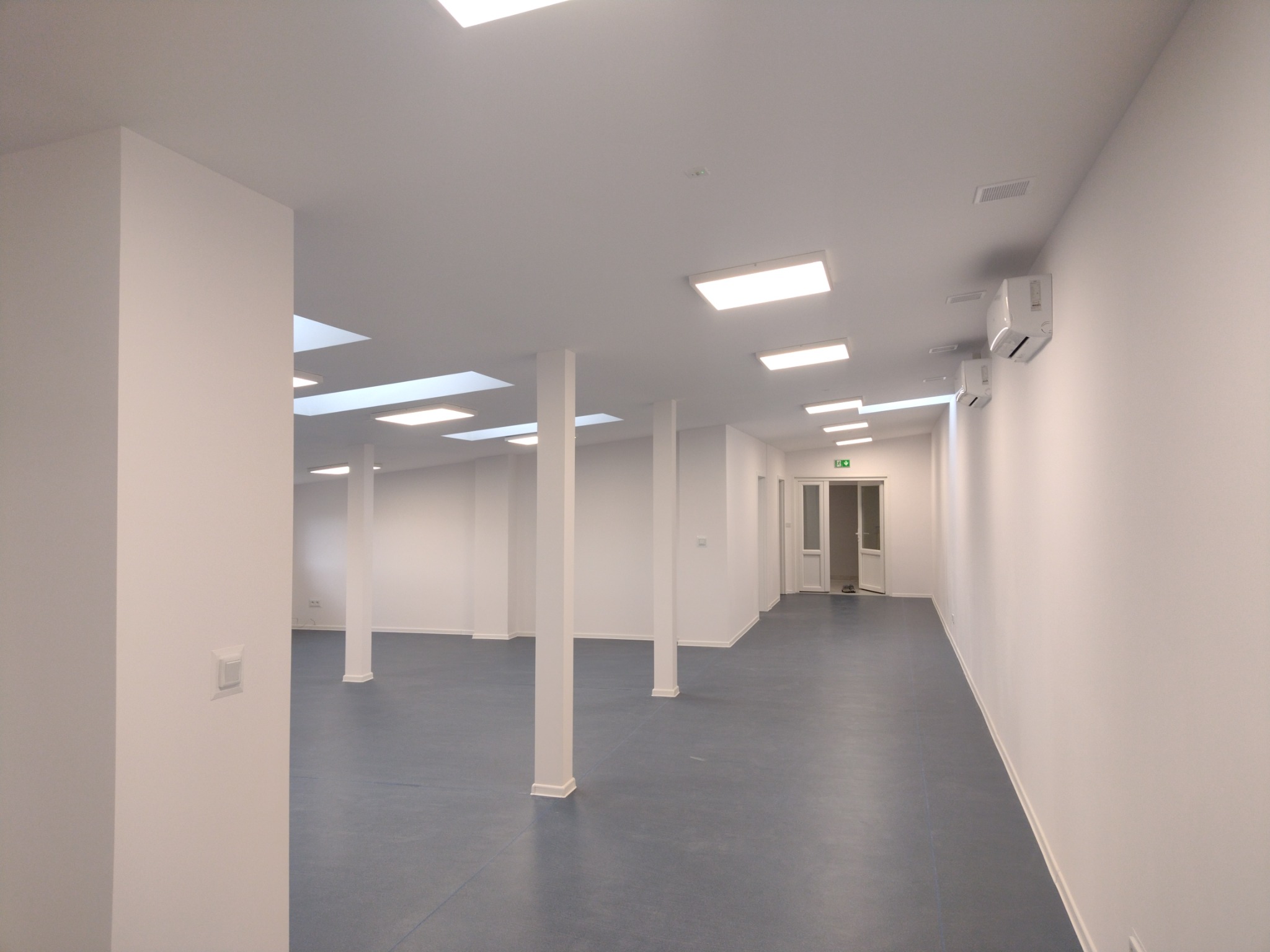 Wnętrze biura po remoncie: białe ściany, niebieska podłoga, kolumny podtrzymujące sufit z oświetleniem panelowym i klimatyzatory na ścianach.