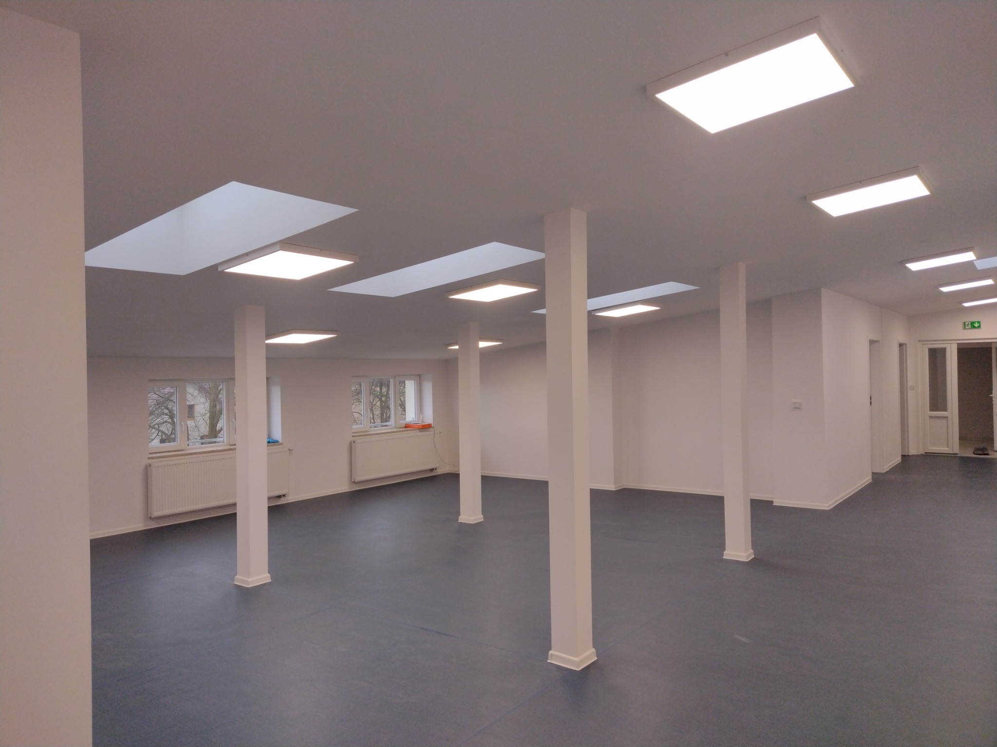 Wnętrze biura po remoncie: białe ściany, szary panel podłogowy, białe kolumny wspierające sufit z wbudowanym oświetleniem rastrowym oraz oknami.