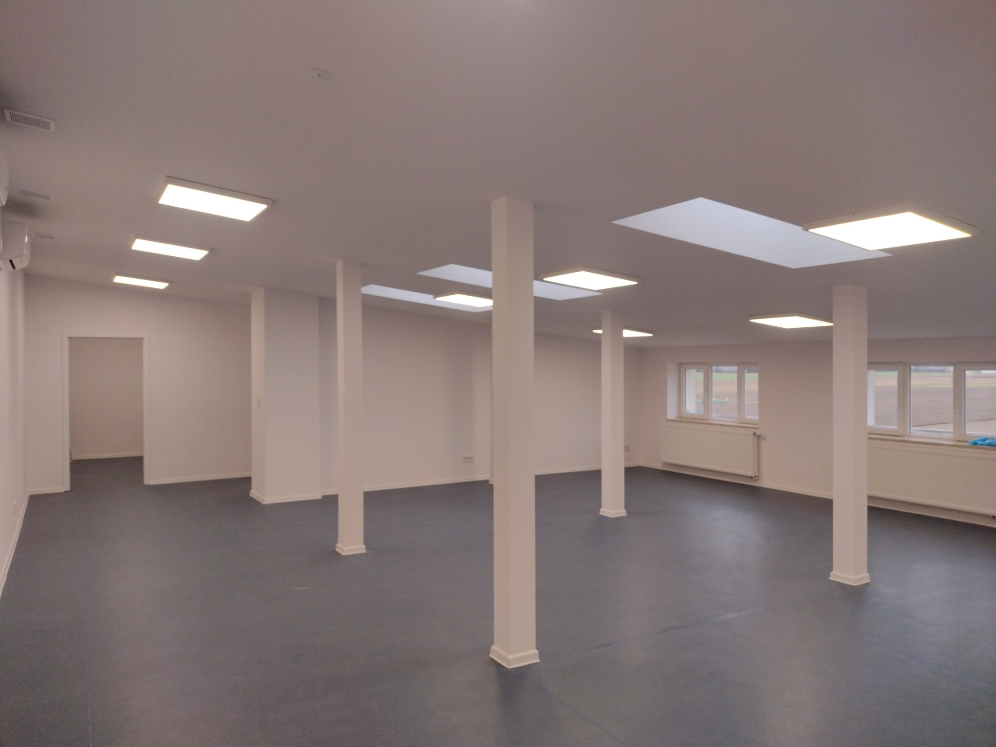 Wnętrze pustego biura po remoncie, z szarą podłogą, białymi ścianami i kolumnami oraz nowoczesnym oświetleniem sufitowym.