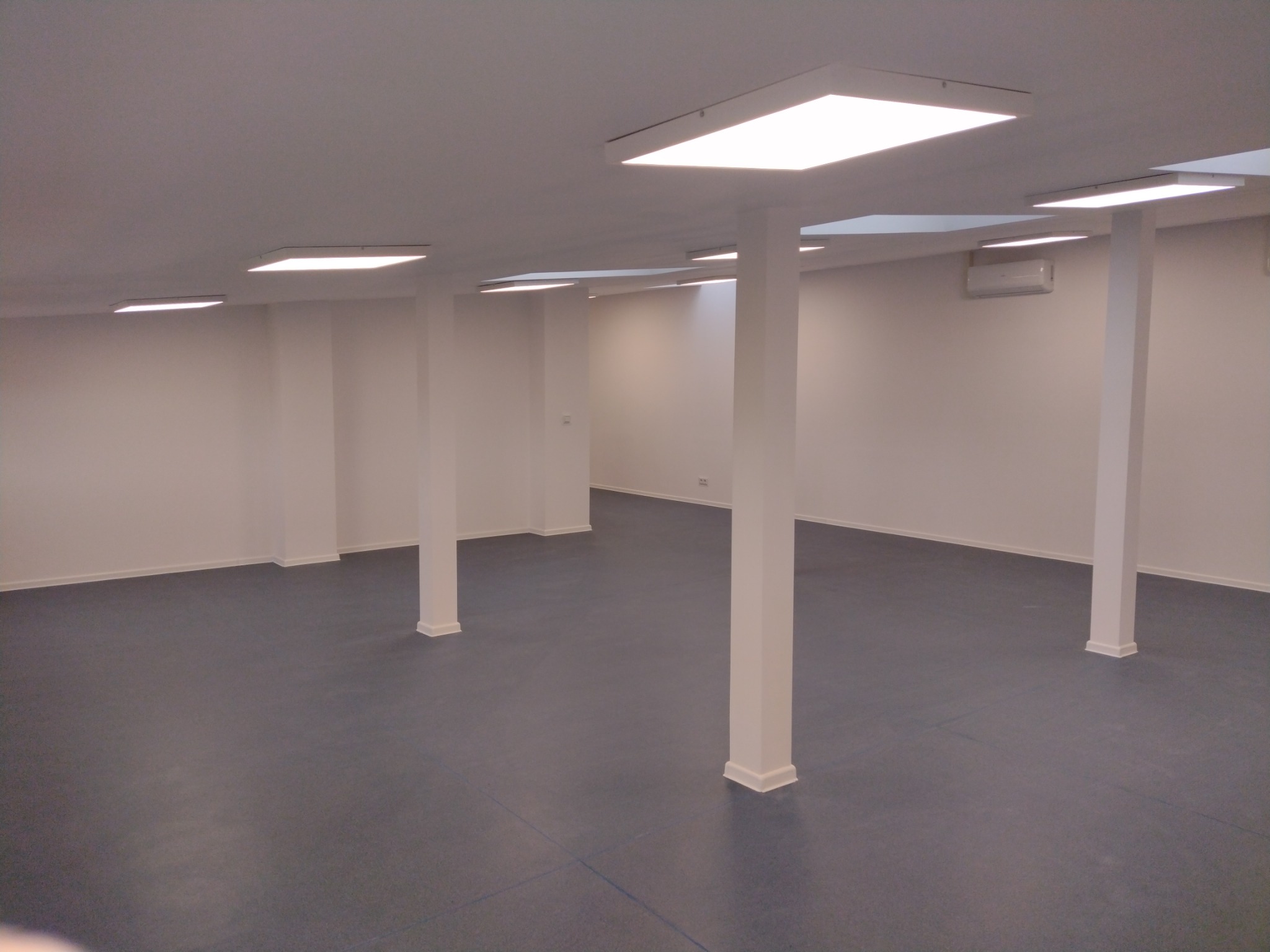 Puste pomieszczenie biurowe z szarym linoleum, białymi ścianami, filarami i oświetleniem sufitowym