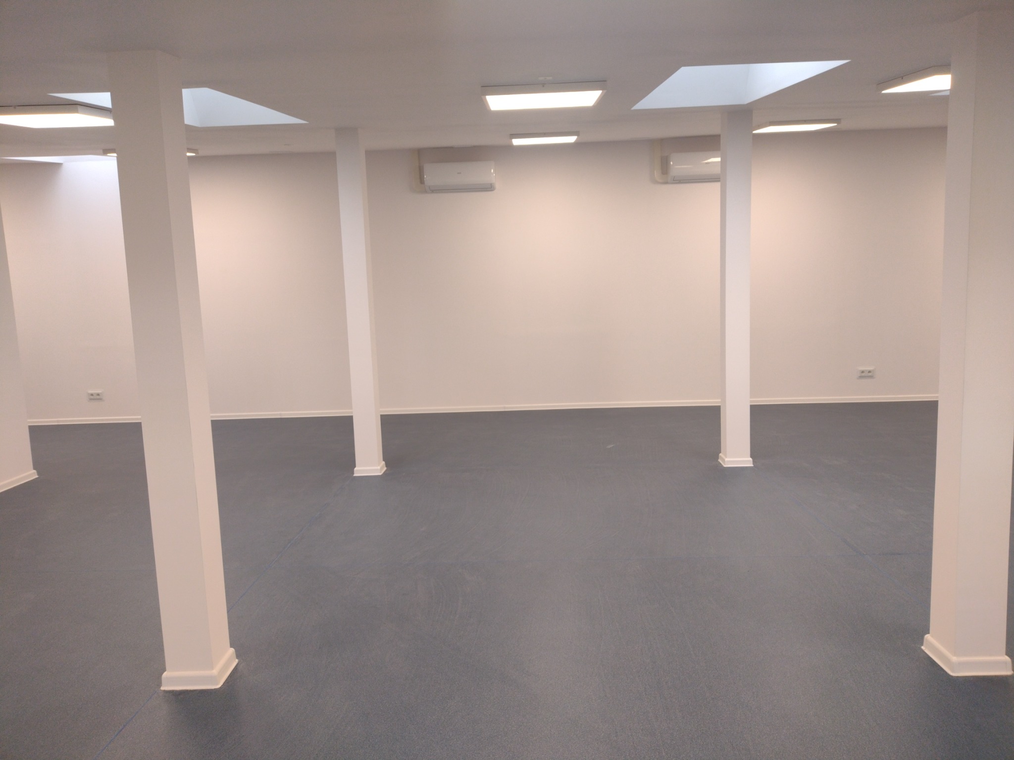 Wnętrze biura po remoncie: szara podłoga, białe ściany z klimatyzatorami, białe kolumny i oświetlenie sufitowe.