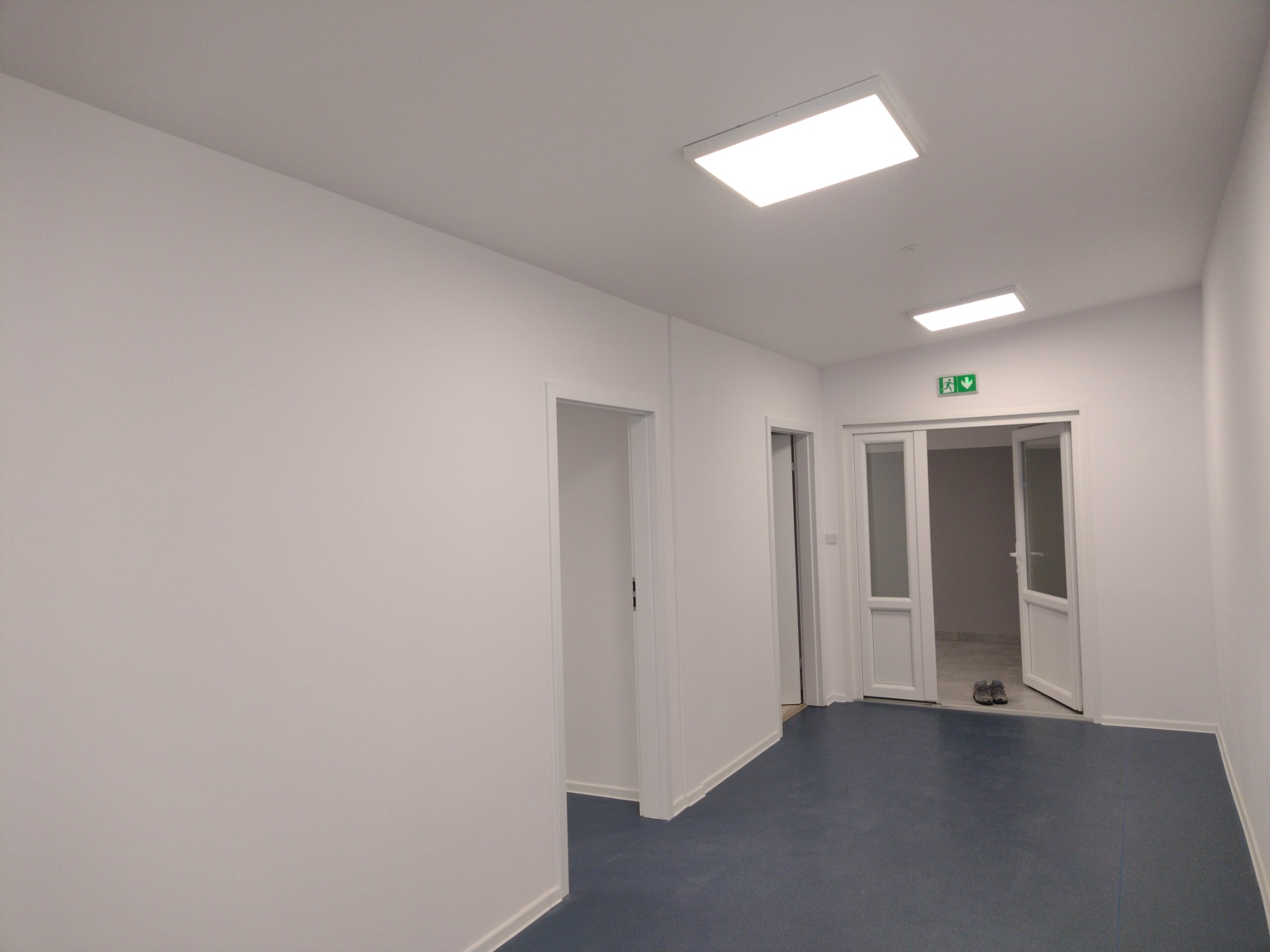 Korytarz biurowy po remoncie z białymi ścianami, niebieską podłogą i widocznymi drzwiami oraz oświetleniem sufitowym.