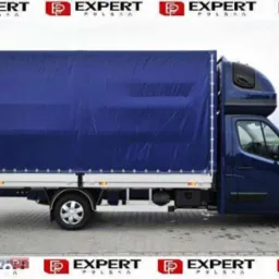 Niebieski bus z plandeką, widok z boku, na aluminiowej platformie, z podwójną kabiną, na tle szarej kostki brukowej i zieleni.