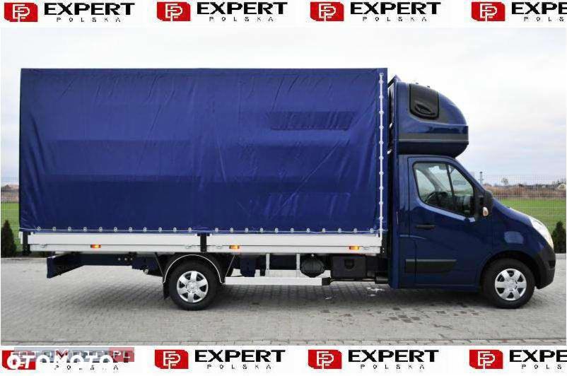 Niebieski bus z plandeką, widok z boku, na aluminiowej platformie, z podwójną kabiną, na tle szarej kostki brukowej i zieleni.