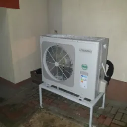 Zewnętrzna jednostka klimatyzacji Hyundai R32 na metalowym stelażu, stojąca na kostce brukowej przy ścianie budynku, lekko przysypana śniegiem.