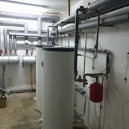 Dwa białe zbiorniki akumulacyjne ciepła w kotłowni, widoczne rury izolowane termicznie, manometry i czerwony zbiornik wyrównawczy, na podłodze ślady wilgoci.