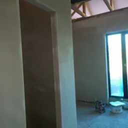 Świeżo nałożona, jasna warstwa tynku na ścianie z widocznym otworem drzwiowym, surowy stan wykończenia wnętrza z widocznymi elementami konstrukcji dachu i oknem w tle.