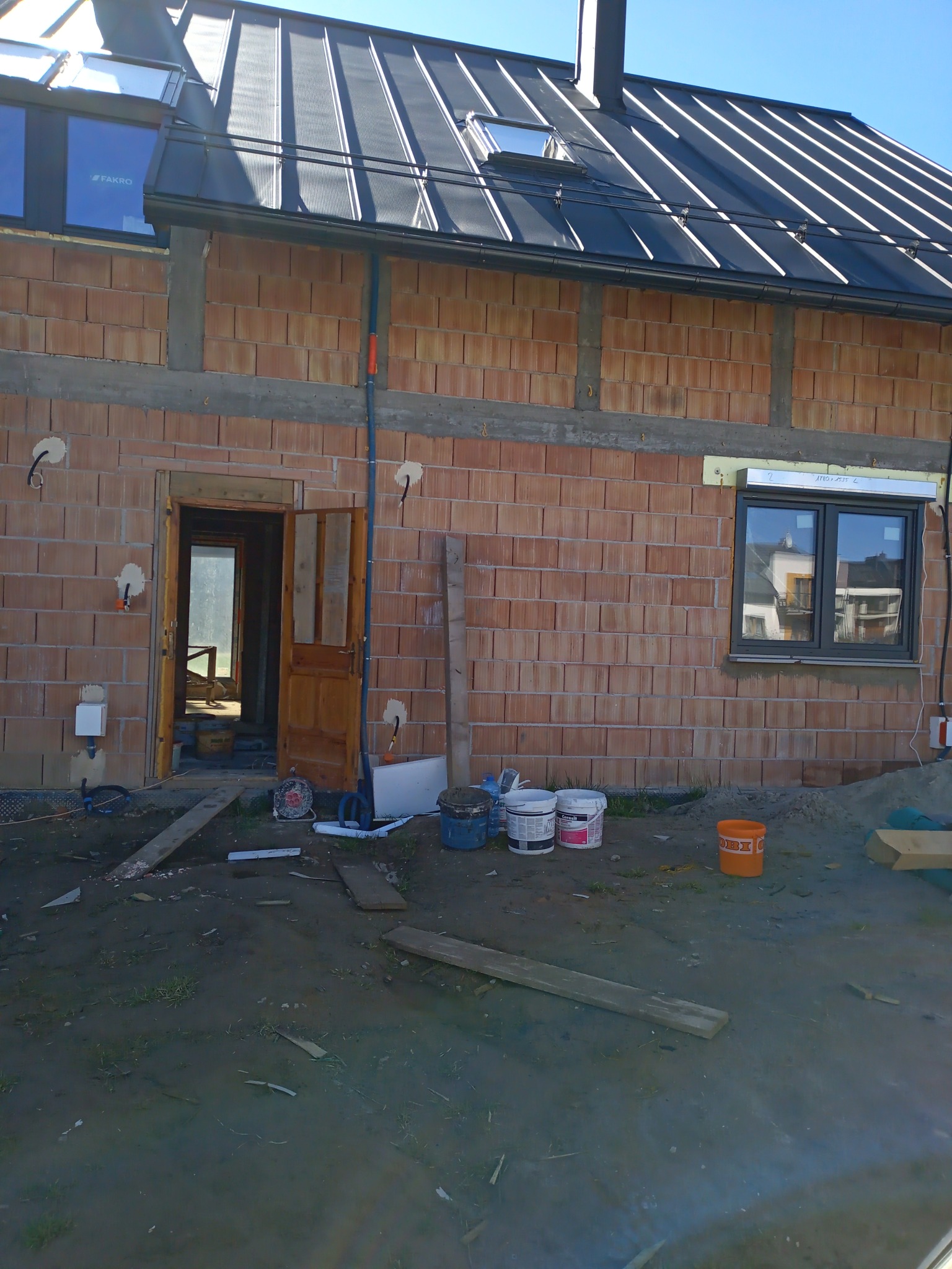 Surowy mur z pustaków ceramicznych, częściowo otynkowany, z otwartymi drzwiami i widocznym wnętrzem, na zewnątrz puste wiadra po zaprawie.