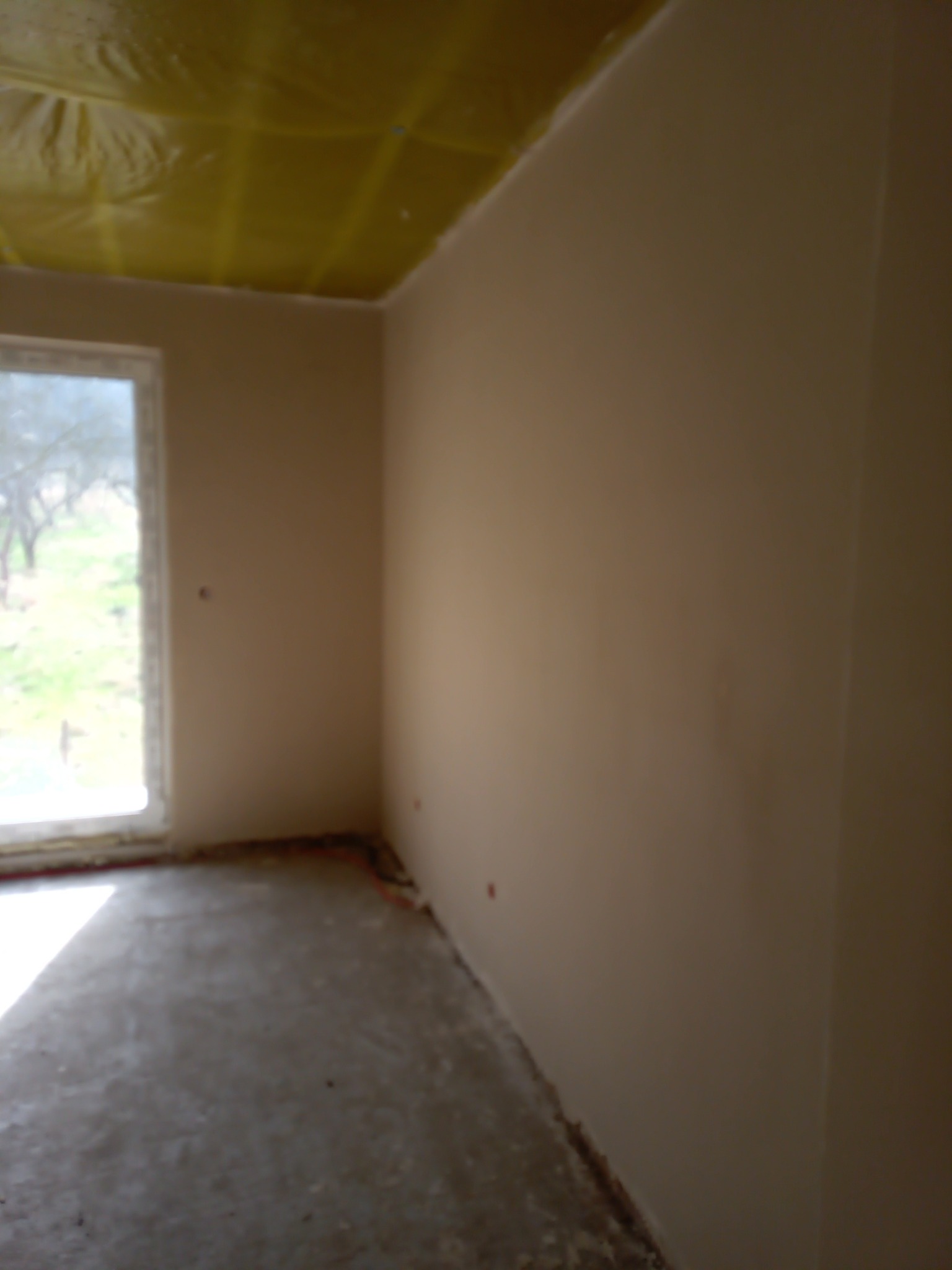 Pokój po nałożeniu gładzi gipsowej, widoczne zabezpieczenie folią malarską sufitu, surowy beton na podłodze i okno z widokiem na zieleń.