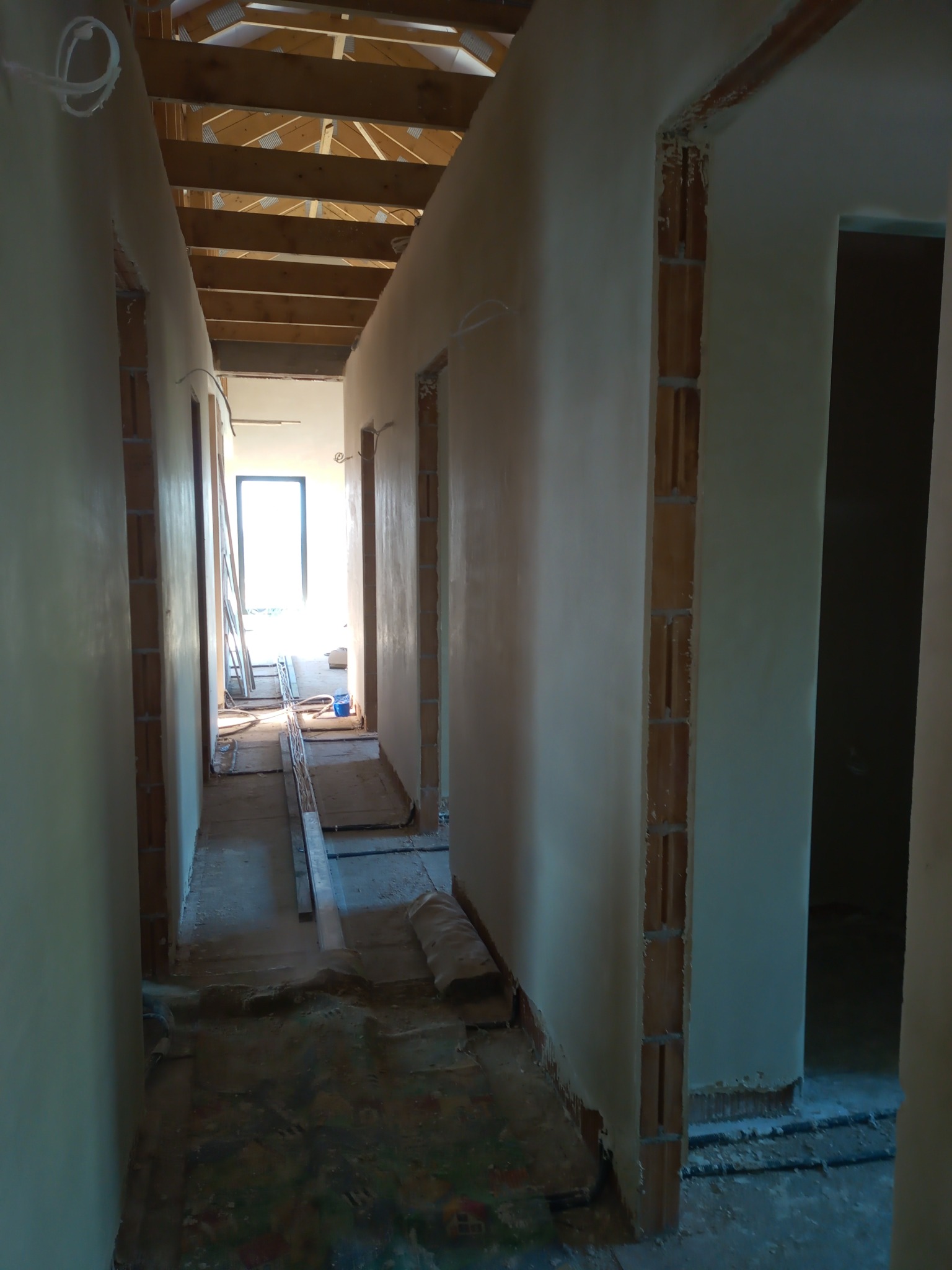 Korytarz w trakcie prac wykończeniowych: ściany pokryte świeżym tynkiem, widoczne elementy konstrukcji drewnianej dachu, podłoga zabezpieczona folią, resztki materiałów budowlanych.