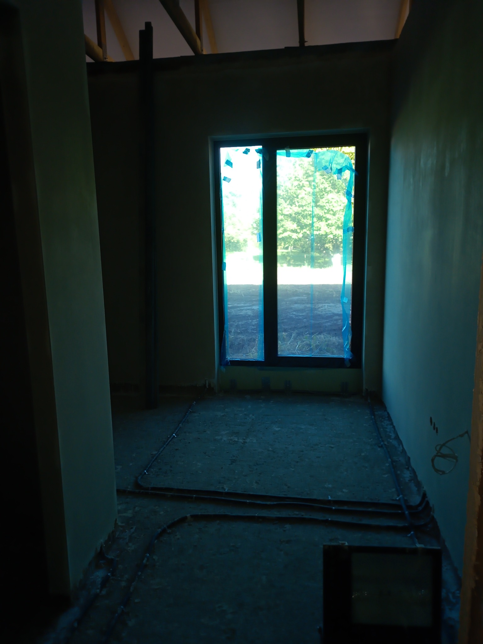 Wnętrze w trakcie prac wykończeniowych: ściany po nałożeniu gładzi, okno zabezpieczone folią, widoczne instalacje elektryczne na podłodze.