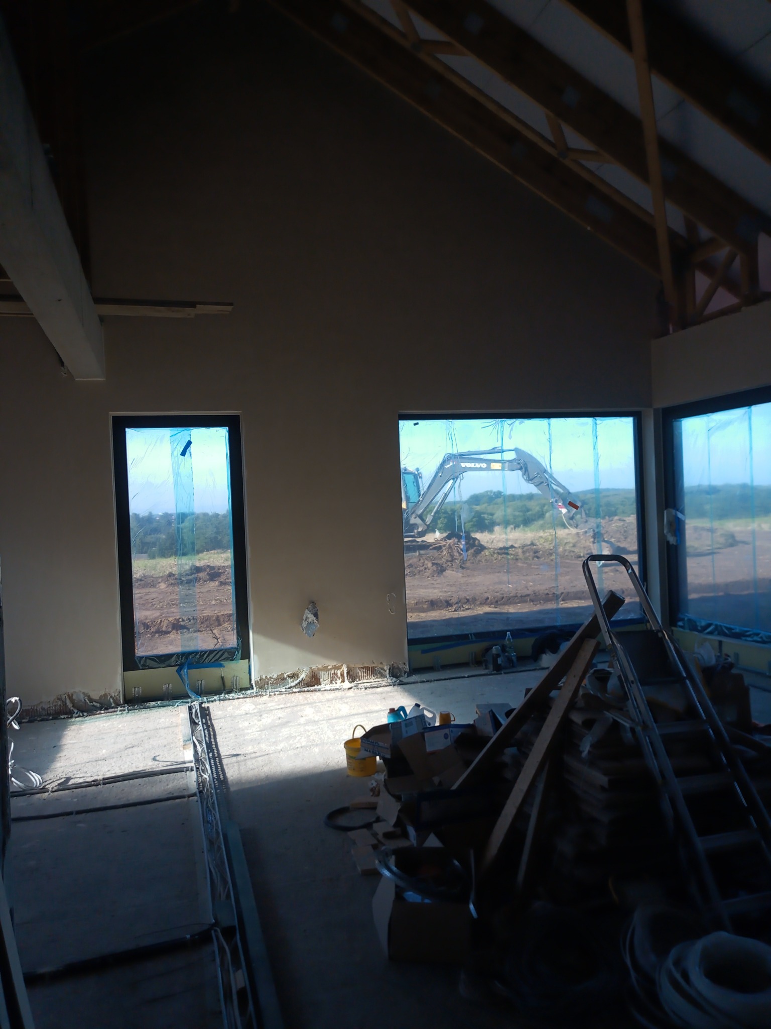 Wnętrze budynku w trakcie prac wykończeniowych: ściany pokryte tynkiem, okna zabezpieczone folią, w tle widoczny sprzęt budowlany na zewnątrz.