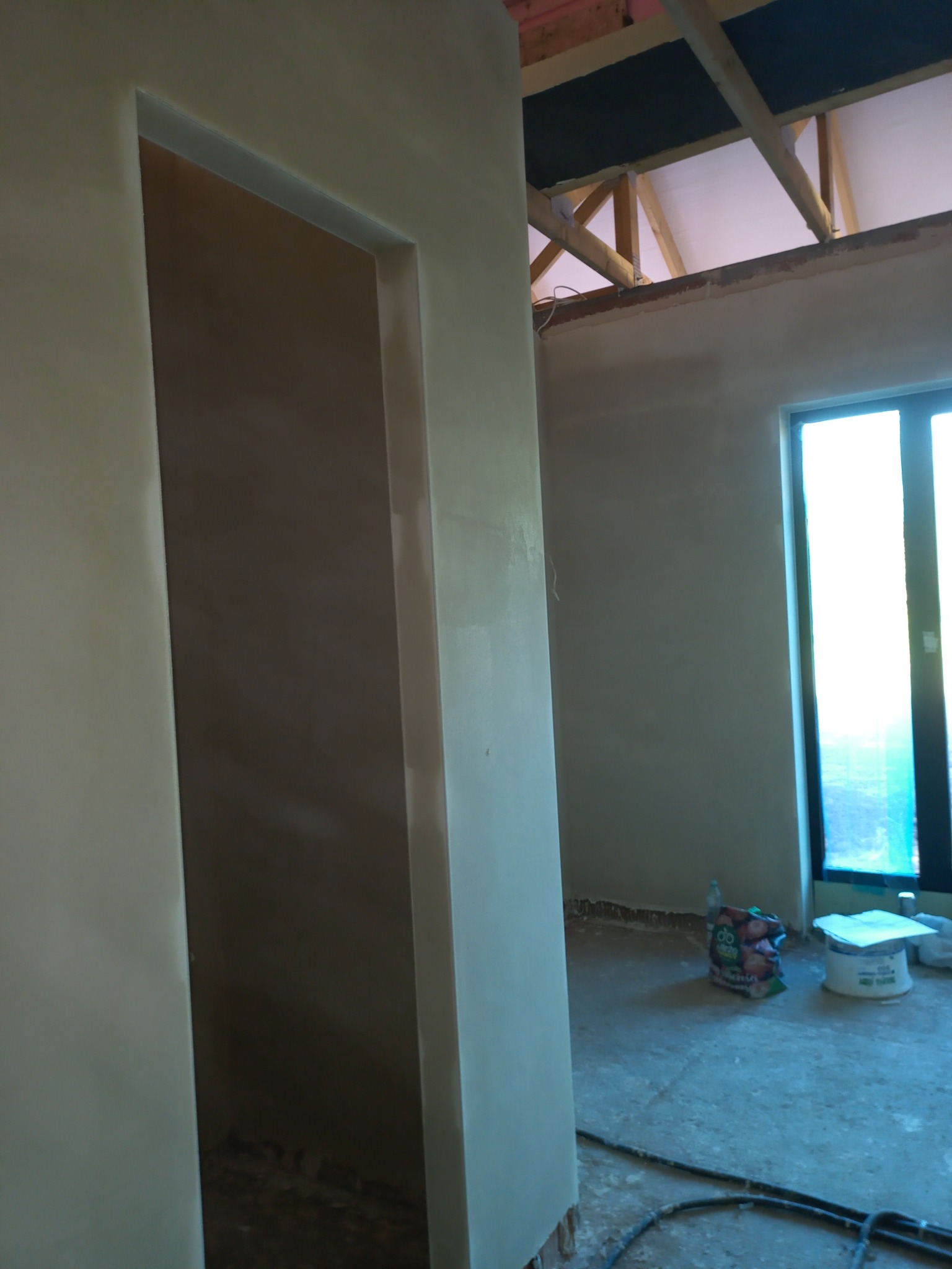 Świeżo nałożona, jasna warstwa tynku na ścianie z widocznym otworem drzwiowym, surowy stan wykończenia wnętrza z widocznymi elementami konstrukcji dachu i oknem w tle.