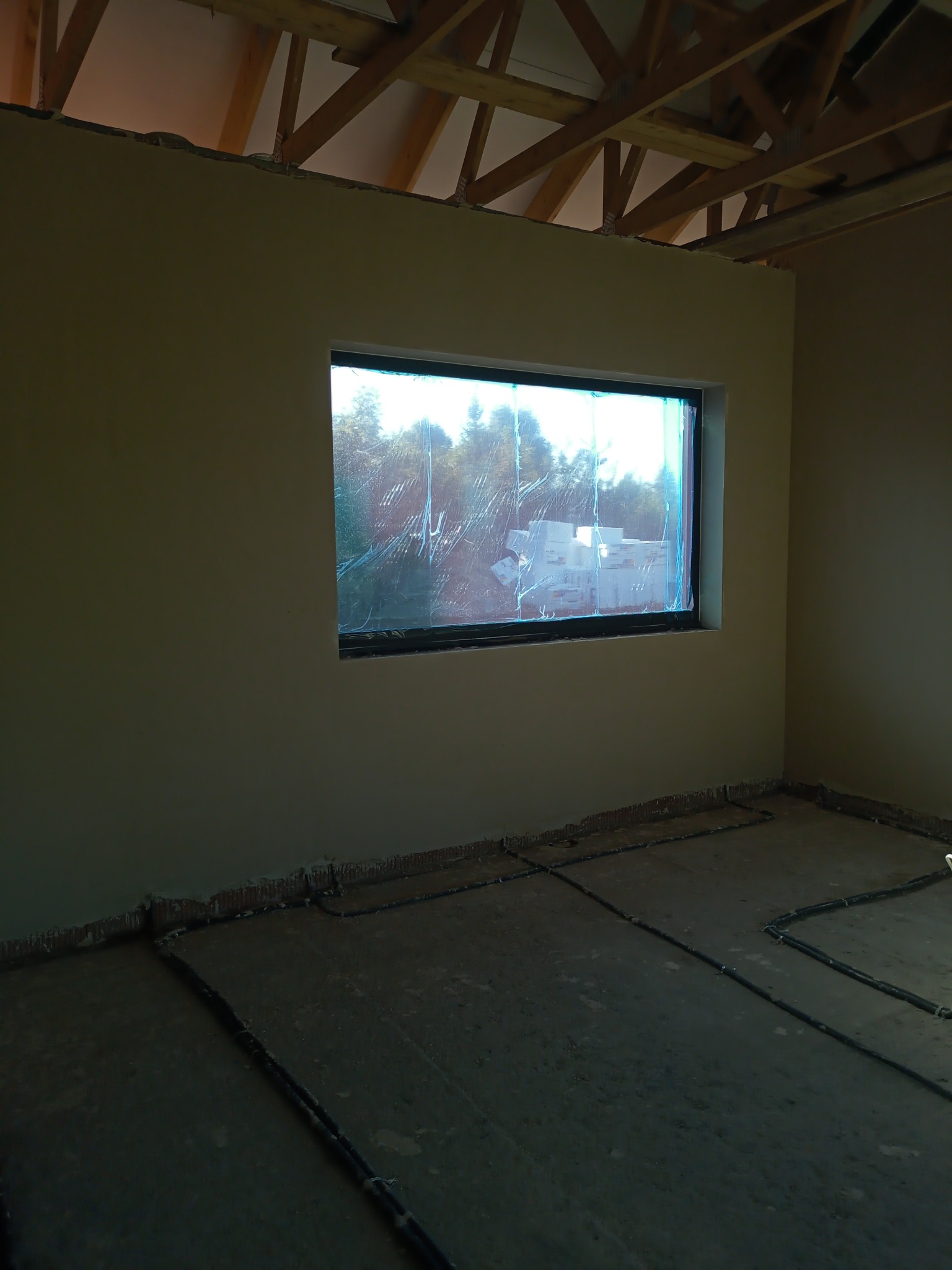 Wnętrze w stanie surowym po nałożeniu gładzi, z widoczną drewnianą konstrukcją dachu, zabezpieczonym oknem folią i instalacją podłogową.