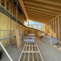 Wnętrze w trakcie budowy: szkielet domu z drewnianych belek, widoczne rusztowanie, izolacja ścian, duże okno z widokiem na zewnątrz i materiały budowlane przygotowane do dalszych prac.