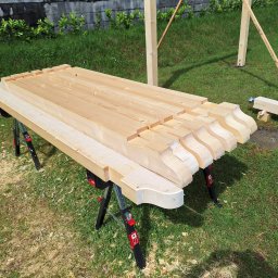 TB Woodhaus - Przygotowane elementy drewniane z ozdobnym wykończeniem, ułożone na stole warsztatowym na trawie, prawdopodobnie do budowy altany lub pergoli.