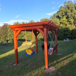 TB Woodhaus - Drewniana konstrukcja z trzema huśtawkami w ogrodzie, w tym jedna plastikowa dla małych dzieci oraz huśtawka typu bocianie gniazdo i jedna klasyczna drewniana, w tle zielona trawa i drzewa.