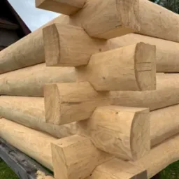 Fragment konstrukcji domu z okrąglaków, widoczne połączenia na węgłach, świeże drewno z widocznymi śladami obróbki, w tle fragment dachu i drzewa.