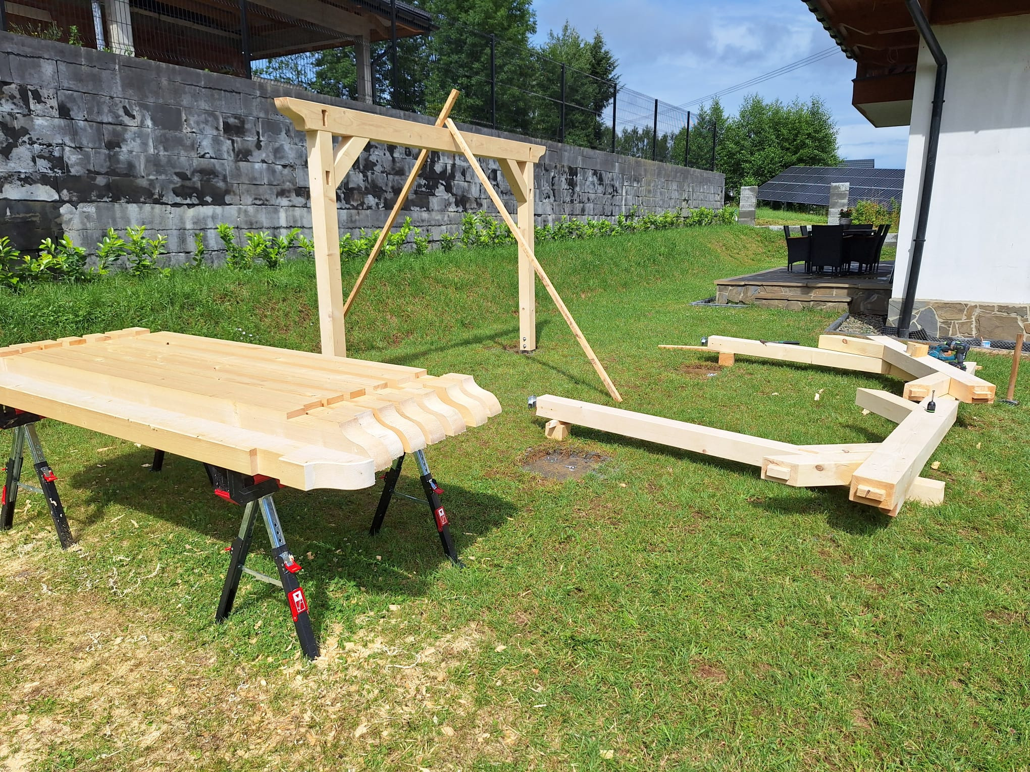 Drewniana konstrukcja w trakcie montażu na trawniku, elementy pergoli rozłożone, widoczne narzędzia i fragment domu w tle.