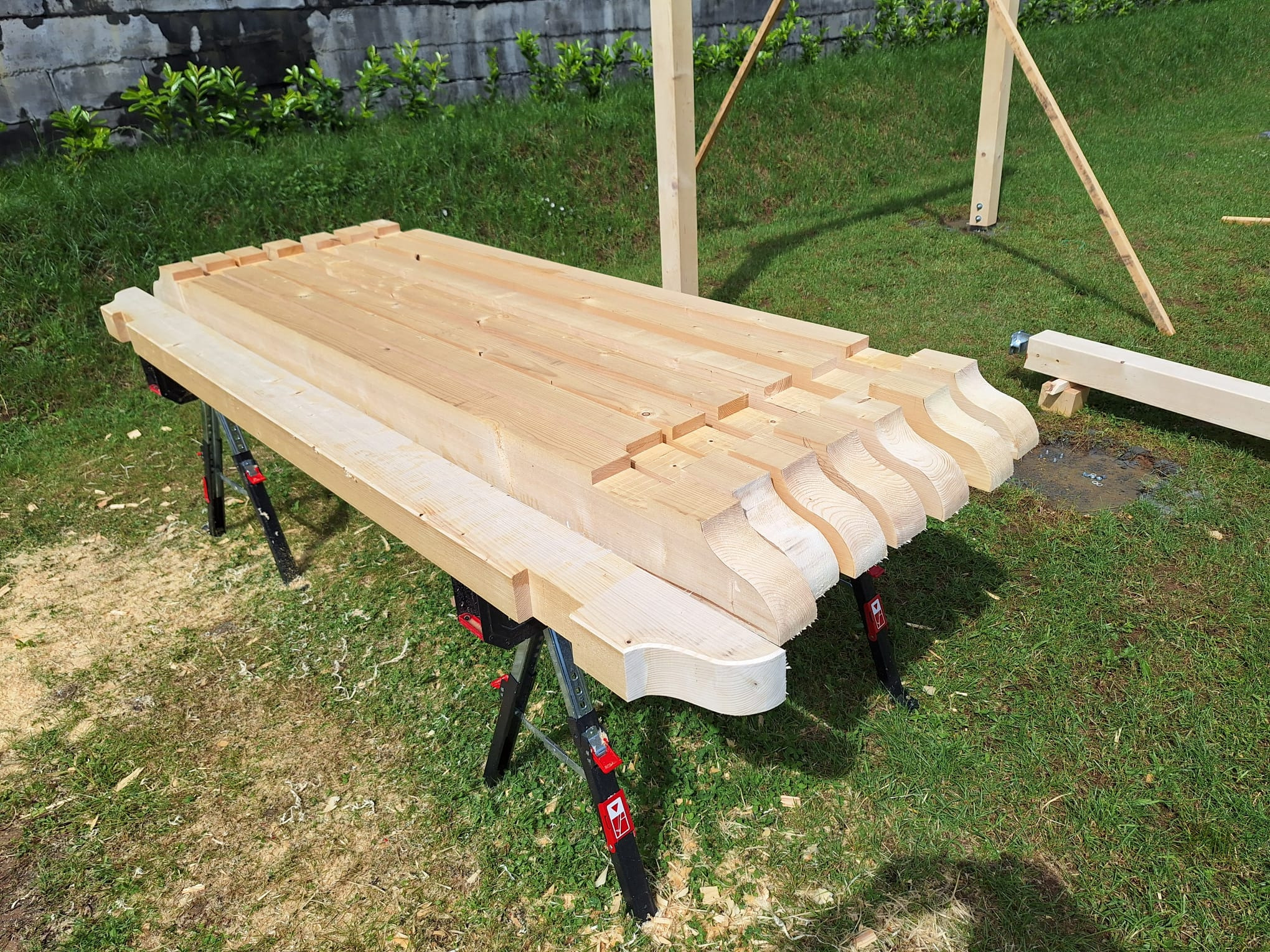 Przygotowane elementy drewniane z ozdobnym wykończeniem, ułożone na stole warsztatowym na trawie, prawdopodobnie do budowy altany lub pergoli.