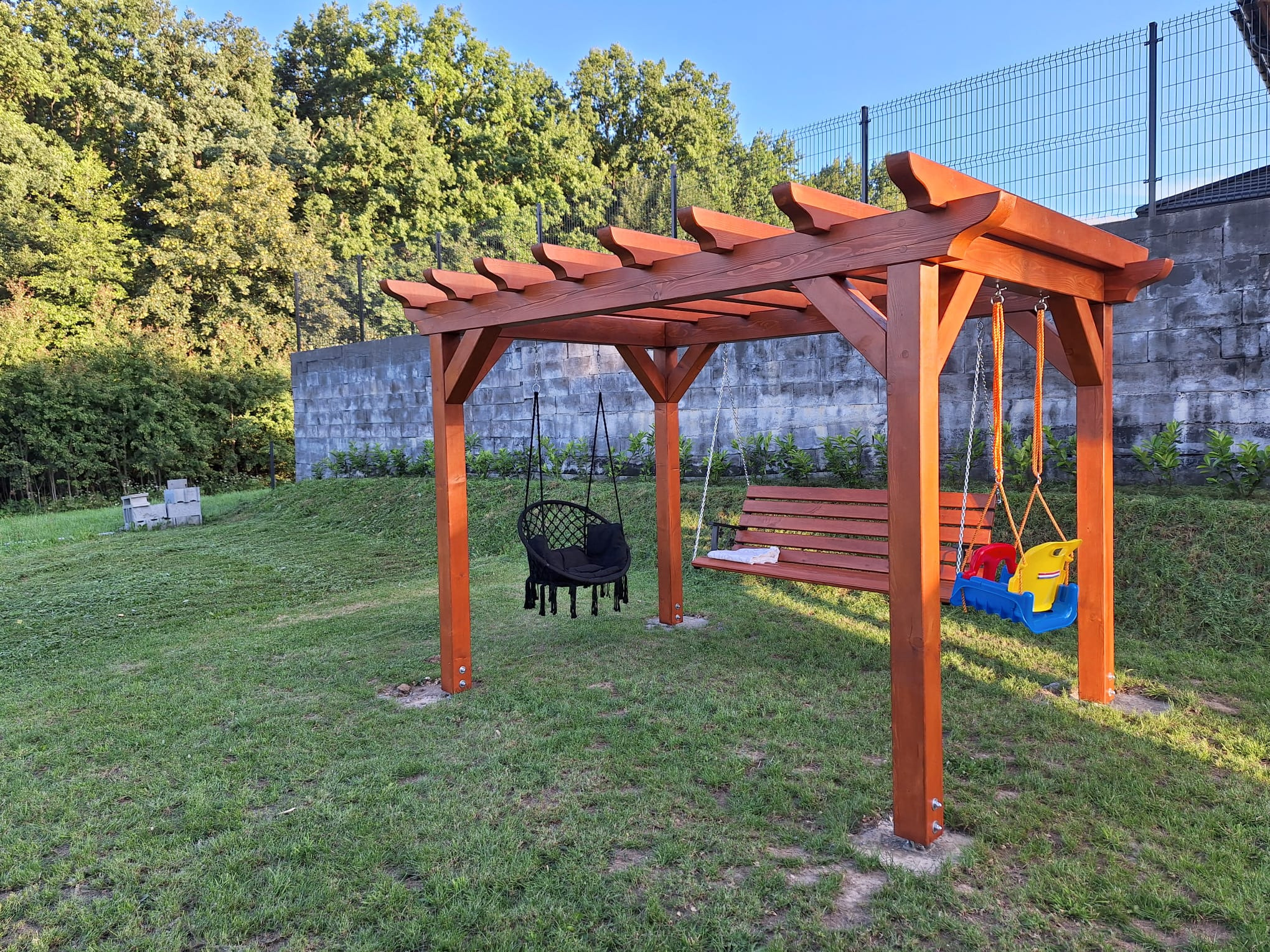 Drewniana konstrukcja z trzema huśtawkami: czarnym fotelem wiszącym, ławką z poduszką i plastikową huśtawką dla dzieci, na trawniku przed murem i zielenią.