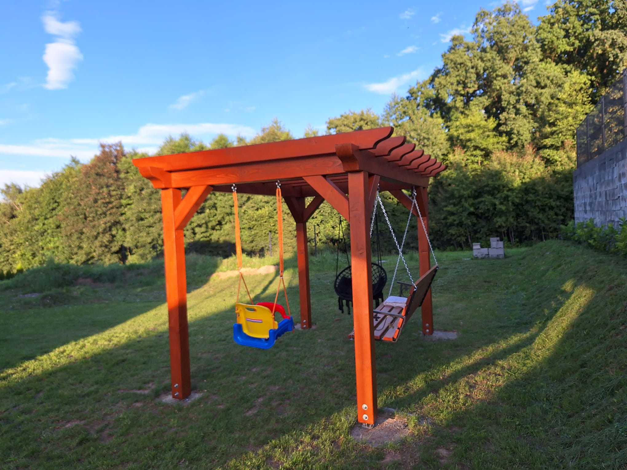 Drewniana konstrukcja z trzema huśtawkami w ogrodzie, w tym jedna plastikowa dla małych dzieci oraz huśtawka typu bocianie gniazdo i jedna klasyczna drewniana, w tle zielona trawa i drzewa.