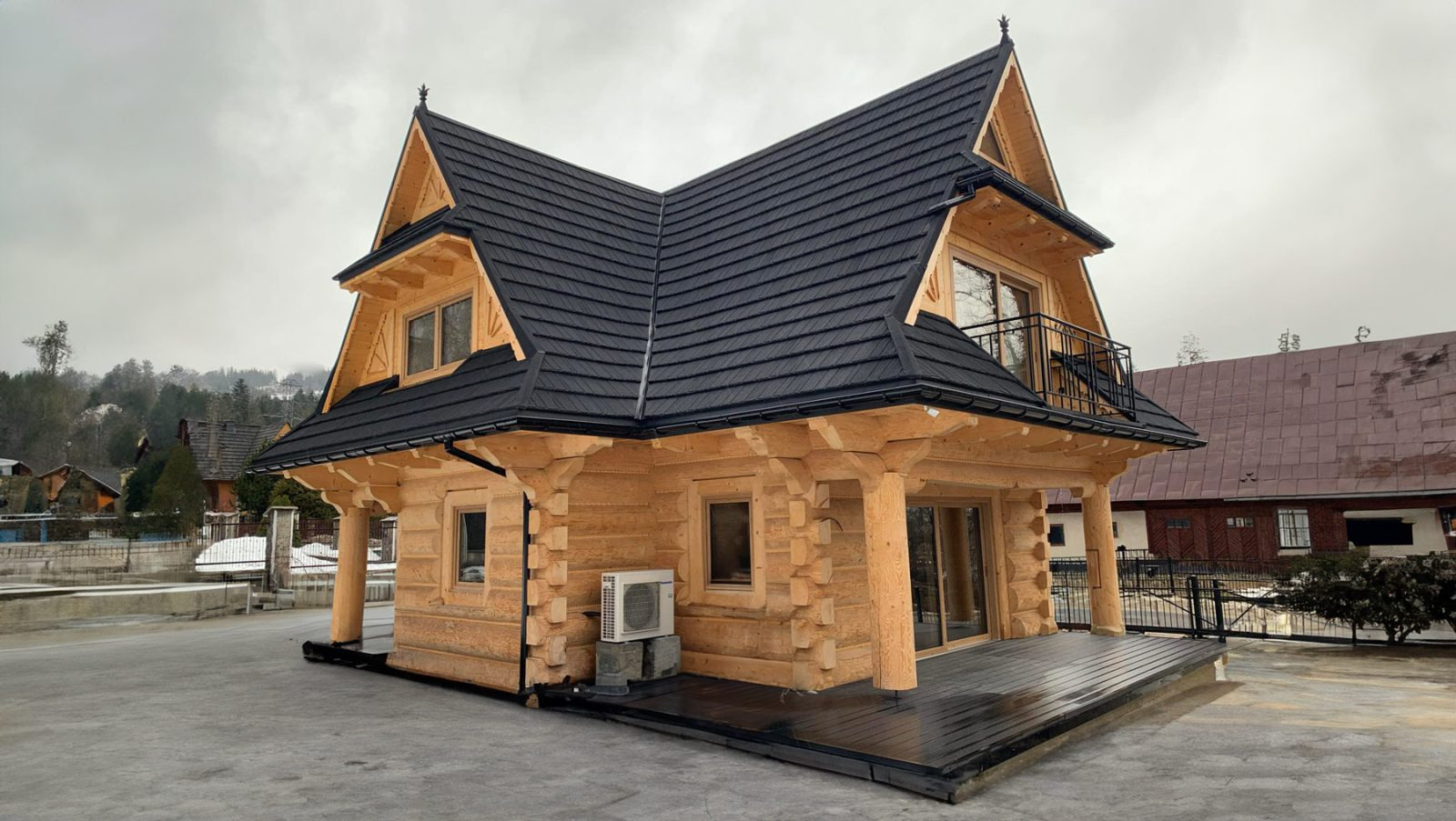 Dwukondygnacyjny dom z drewnianych bali z czarnym dachem, balkonem i klimatyzatorem zewnętrznym, na tle zabudowy i zalesionego wzgórza w pochmurny dzień.