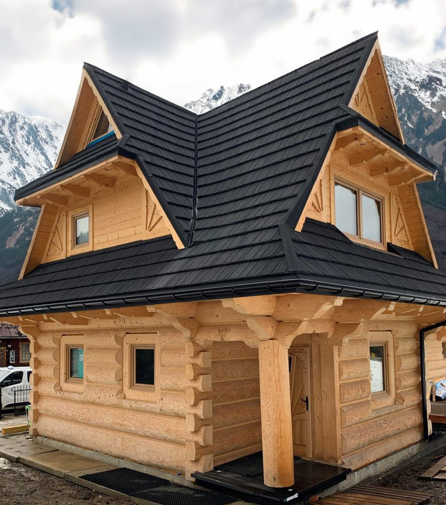 Nowoczesny, parterowy budynek z bali, z czarnym dachem z blachodachówki na tle górskiego krajobrazu, widoczne detale wykończenia i drewniana konstrukcja.