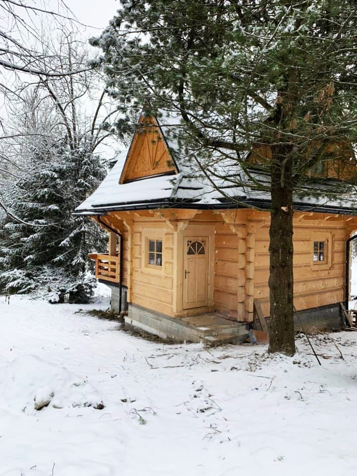 Mały, parterowy budynek z drewna, przykryty śniegiem, z widocznym drewnianym balkonem i schodami wejściowymi, otoczony zimowym krajobrazem z drzewami pokrytymi śniegiem.