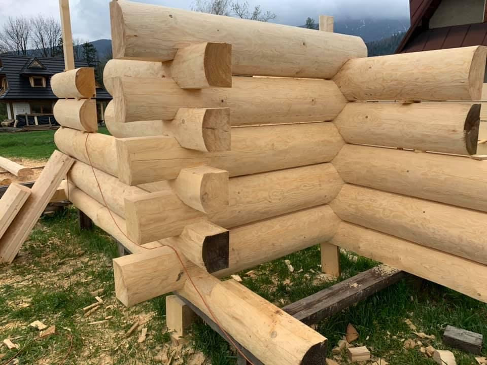 Fragment budowy domu z bali, widoczne obrobione bale z charakterystycznymi wycięciami na rogach, ułożone warstwami, z widocznym tłem górskim i częścią innego budynku.
