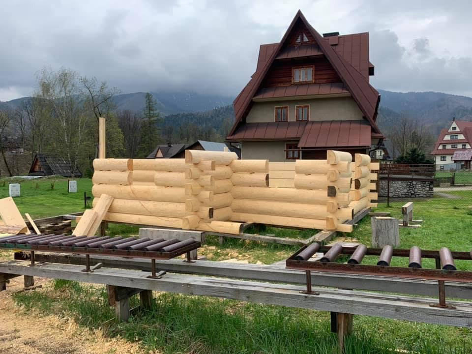 Budowa domu z okrąglaków na fundamencie, widoczne podkłady rolkowe, w tle zabudowa jednorodzinna i zalesione wzgórza pod pochmurnym niebem.