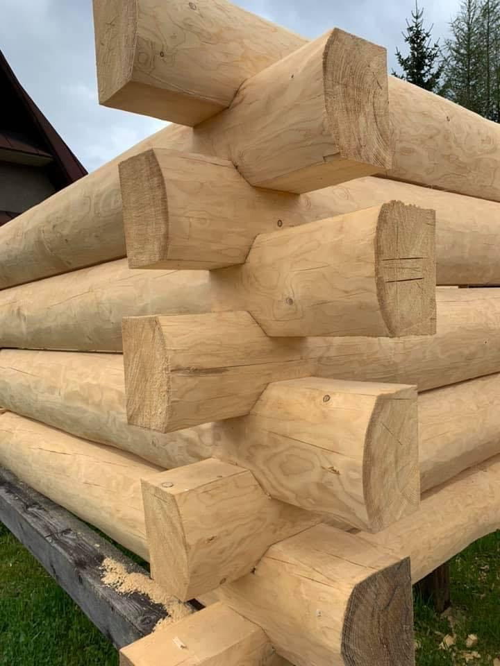 Fragment konstrukcji domu z okrąglaków, widoczne połączenia na węgłach, świeże drewno z widocznymi śladami obróbki, w tle fragment dachu i drzewa.