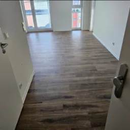 Nowoczesne wnętrze z ciemną, drewnopodobną podłogą ułożoną w panelach, widoczne okna z siatką zabezpieczającą i fragment otwartych drzwi z klamką.