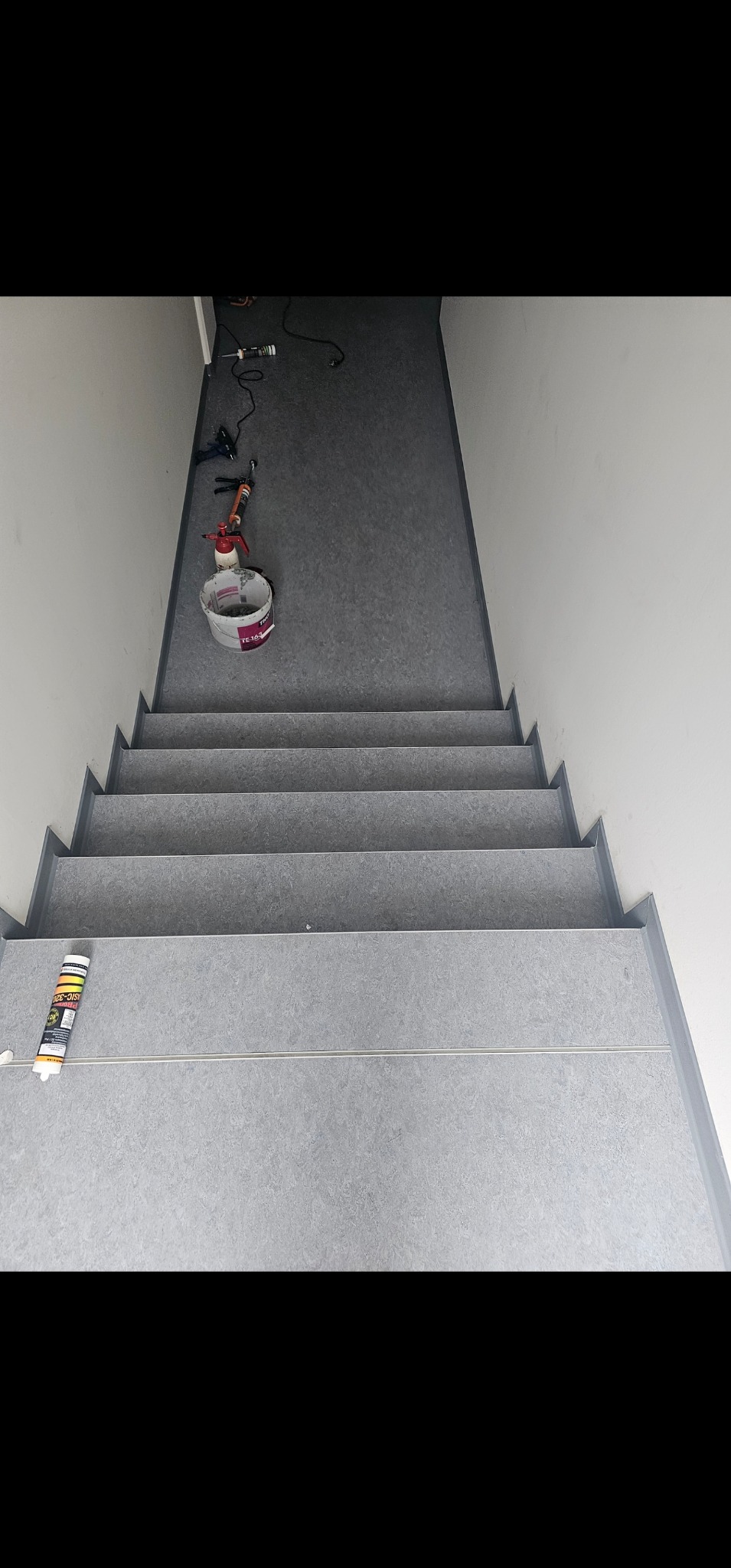 Szare schody wyłożone szarą wykładziną, widok z góry, z narzędziami i wiadrem z klejem na górnym podeście.