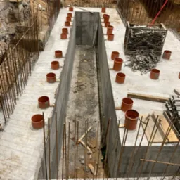 Wykonany fundament szczelinowy z wystającymi prętami zbrojeniowymi i pomarańczowymi rurami osłonowymi, widoczne elementy szalunkowe i grunt.