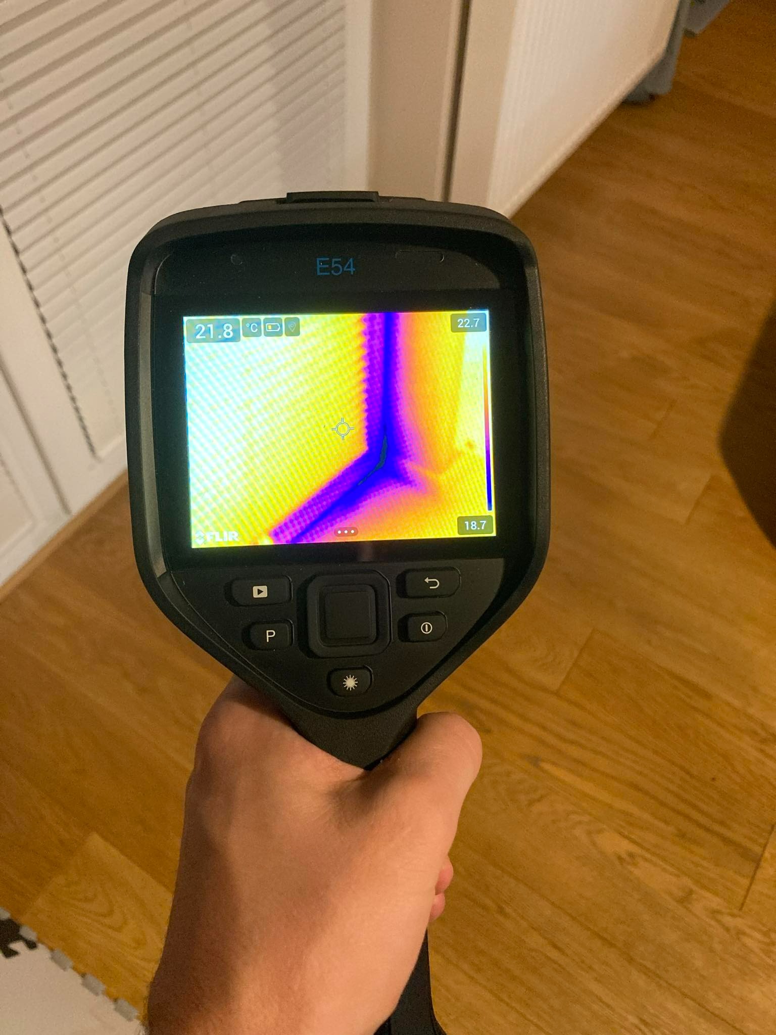 Ręka trzymająca kamerę termowizyjną FLIR E54, wyświetlającą obraz termiczny rogu ściany z widocznymi różnicami temperatur oznaczonymi kolorami na tle drewnianej podłogi.