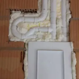 Instalacja rur systemu centralnego odkurzania w ścianie z cegły, zabezpieczona pianką montażową i osłonięta panelem.