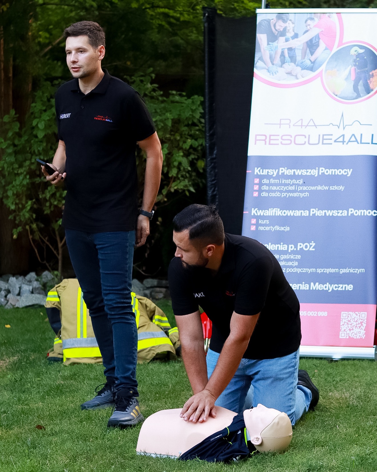 Instruktor demonstruje resuscytację krążeniowo-oddechową na manekinie podczas szkolenia, drugi instruktor obserwuje z telefonem w dłoni, w tle baner informacyjny i ubranie strażackie.