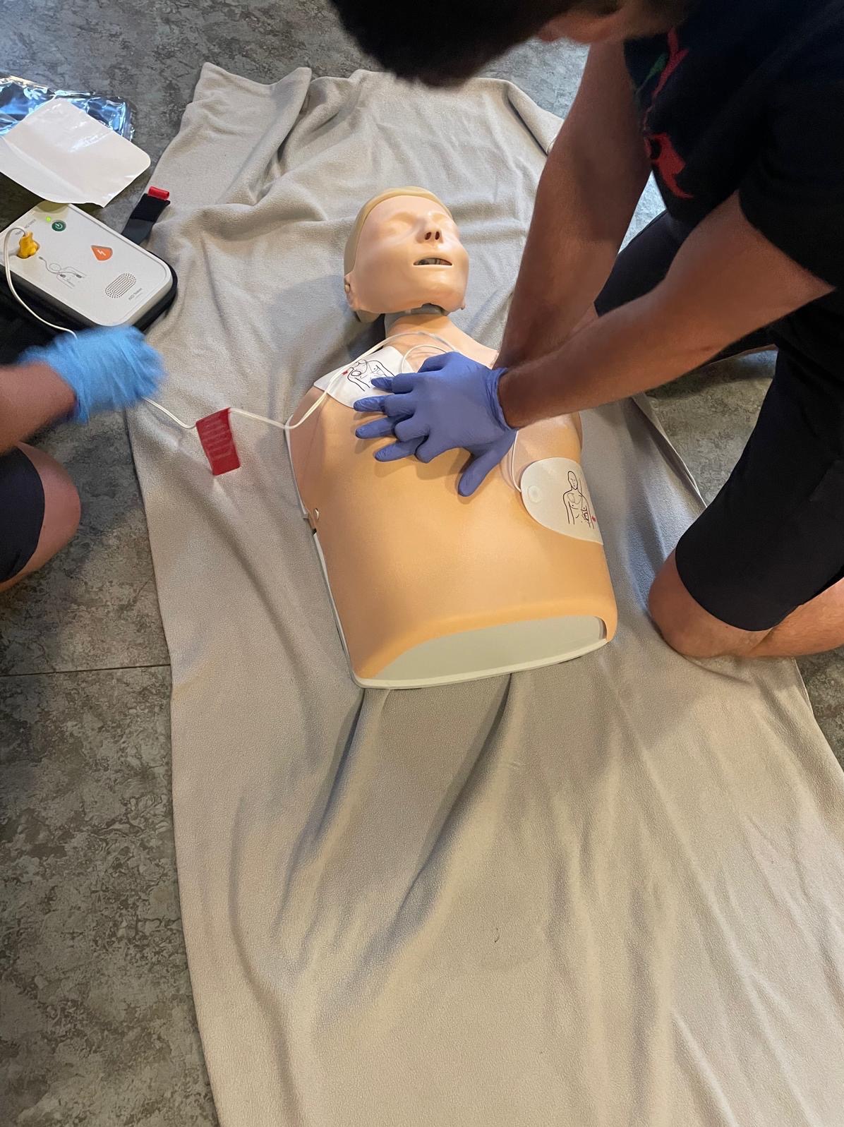 Instruktorzy w niebieskich rękawiczkach demonstrujący uciskanie klatki piersiowej na manekinie podczas kursu pierwszej pomocy z użyciem defibrylatora AED.