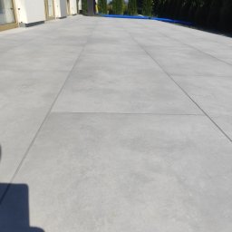 MR-BUD Patryk Mróz - Nowoczesny taras wyłożony dużymi, szarymi płytami. Widoczne geometryczne linie fug, budynek w tle i fragment niebieskiego węża ogrodowego.