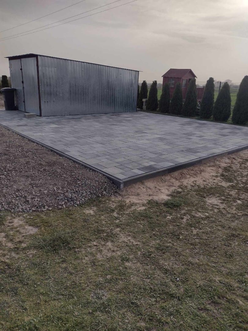 Ułożona szara kostka brukowa tworząca plac przed blaszanym garażem, obrzeża z ciemnej kostki, w tle rząd tuj i mały drewniany domek.