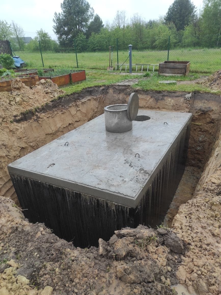 Betonowy zbiornik, prawdopodobnie szambo, umieszczony w wykopie na działce z widocznym ogrodzeniem i zielenią w tle.