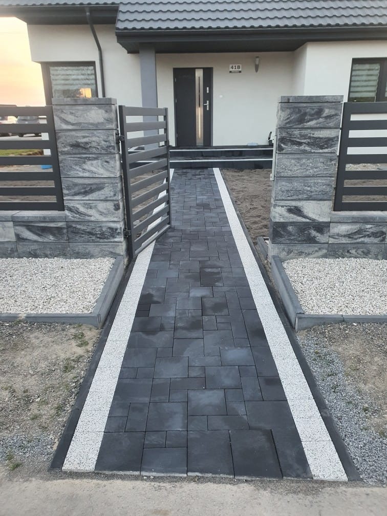 Nowoczesny podjazd z ciemnej kostki brukowej, obramowany białym grysem, prowadzący do domu z szarą stolarką okienną i otwartą furtką.