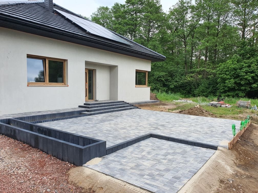 Ułożona kostka brukowa na posesji domu jednorodzinnego z panelami słonecznymi na dachu, widoczne obrzeża i krawężniki, w tle las.