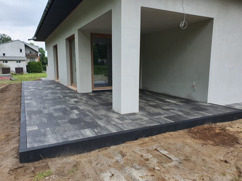 Nowoczesny taras z szarej kostki brukowej ułożonej przy białym budynku z dużymi oknami, obrzeża tarasu wykończone czarną kostką.