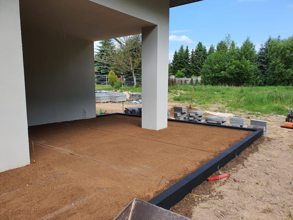 Przygotowany teren pod wiatą z obrzeżami, zagęszczona podsypka, palety z kostką w tle, widoczna zagęszczarka.